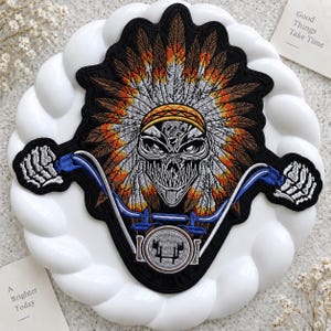 May include: An embroidered patch featuring a skull wearing a large feathered headdress in shades of orange, brown, and grey. The skull is positioned behind blue motorcycle handlebars with skeletal hands gripping the ends and a circular headlight below.