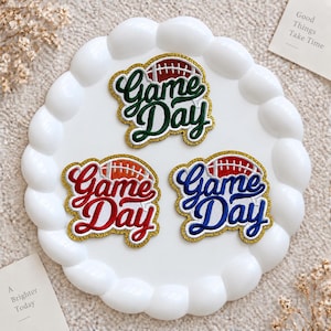 Puede incluir: Tres parches bordados termoadhesivos con el texto Game Day en letra cursiva. Cada parche incluye un gráfico de balón de fútbol americano y un borde de purpurina dorada. Los parches son de color verde oscuro, rojo brillante y azul real.