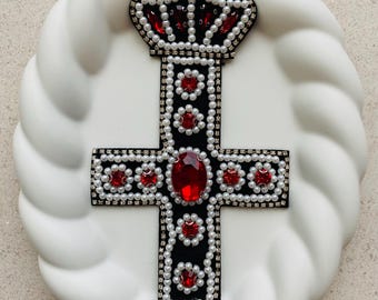 May include: A decorative cross with a crown, featuring a black base adorned with red jewels and faux pearls. The cross is centered on a white, textured surface, creating a striking contrast. The crown is at the top of the cross.