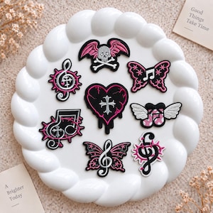 Puede incluir: Una colección de parches bordados con diseños en negro, blanco y rosa que incluyen una calavera con alas de murciélago, un corazón con una cruz, mariposas, notas musicales y claves de sol, dispuestos sobre un plato blanco circular festoneado.
