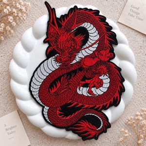 Pode incluir: Um emblema bordado apresentando um dragão detalhado em vermelho e preto com a barriga branca. O dragão é retratado em uma posição enrolada com garras afiadas e escamas complexas. O design é contornado com fio preto, criando um forte contraste.