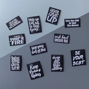 May include: A collection of black embroidered patches with white text. The patches feature motivational phrases like "Never Give Up," "Be Your Best," and "Work Hard Dream Big." The patches are rectangular and have a white border.
