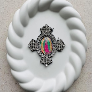 May include: A decorative cross featuring a central image of Our Lady of Guadalupe, surrounded by sparkling rhinestones. The cross is set against a white, textured, oval-shaped plate. The image within the cross showcases vibrant colors.
