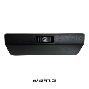 Peut inclure: Boîte à gants de voiture noire avec une surface texturée. La boîte à gants est de forme rectangulaire avec un loquet portant l'inscription "WOLFSBURG EDITION" et un emblème rouge et blanc. Le texte "GOLF MK2 PARTS . COM" est en bas.