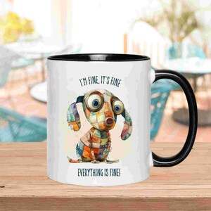 May include: White ceramic mug with a black interior and handle. The mug features a whimsical illustration of a patchwork dog with large eyes and the text "I'M FINE, IT'S FINE" and "EVERYTHING IS FINE!"