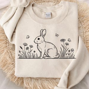 May include: Cream-colored sweatshirt featuring a black line drawing of a rabbit sitting among flowers and butterflies. The design is centered on the front of the sweatshirt. The sweatshirt has a crew neck and long sleeves.