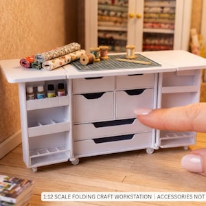 Puede incluir: Una mesa de manualidades plegable blanca a escala 1:12 con múltiples cajones, estantes laterales y una alfombrilla de corte verde en la superficie. Incluye bobinas de madera, rollos de tela y frascos. Texto: 1:12 SCALE FOLDING CRAFT WORKSTATION.