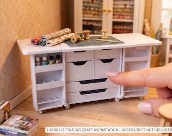 May include: A white 1:12 scale folding craft workstation featuring multiple drawers, side storage shelves, and a flat top surface. The unit includes a green cutting mat, small wooden spools, and fabric rolls. Text reads: 1:12 SCALE FOLDING CRAFT WORKSTATION | ACCESSORIES NOT INCLUDED.