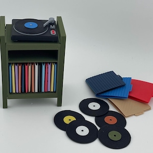 Pu&ograve; includere: Un supporto in miniatura per giradischi color verde oliva con un giradischi nero sopra e uno scaffale pieno di copertine colorate per dischi. Diversi dischi in vinile neri sciolti con etichette centrali colorate e tre custodie quadrate sono vicini.