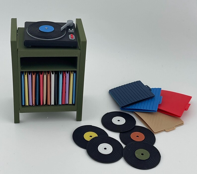 Pu&ograve; includere: Un supporto in miniatura per giradischi color verde oliva con un giradischi nero sopra e uno scaffale pieno di copertine colorate per dischi. Diversi dischi in vinile neri sciolti con etichette centrali colorate e tre custodie quadrate sono vicini.