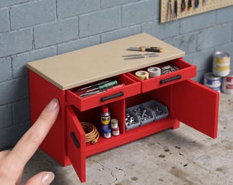 May include: A 1:12 scale red workbench with a beige top, two drawers, and two cabinet doors. The unit features black handles and contains miniature tools, hardware bins, and supplies. The text 1:12 SCALE WORKBENCH | ACCESSORIES NOT INCLUDED is visible.