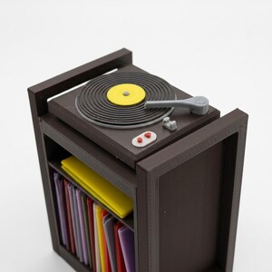 Pu&ograve; includere: Una console giradischi in miniatura marrone con un piatto nero, un'etichetta centrale gialla e un braccio grigio. Sotto il piatto, un ripiano contiene una collezione di sottili copertine per dischi multicolori.