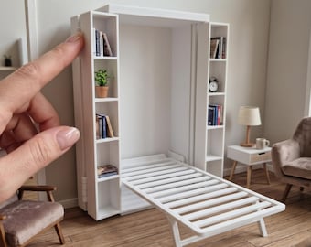 May include: A white 1:12 scale Murphy bed featuring integrated vertical shelving units on both sides. The bed frame is in a lowered, horizontal position with a slatted base. The text 1:12 SCALE MURPHY BED - FULLY FUNCTIONAL | ACCESSORIES NOT INCLUDED is visible.
