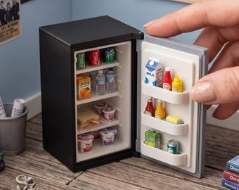 May include: A miniature black refrigerator with a white interior, featuring shelves stocked with tiny food and drink items including juice cartons, soda cans, water bottles, yogurt cups, and a sandwich. The door shelves hold milk, condiments, and butter.