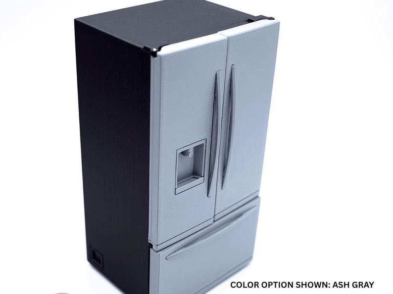 May include: A miniature refrigerator with a black exterior frame and ash gray doors. The unit features two vertical doors with handles, a built-in water dispenser on the left door, and a bottom freezer drawer. The text reads: COLOR OPTION SHOWN: ASH GRAY.