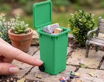 May include: A miniature green wheeled bin in 1:12 scale with an open lid. The bin features a rectangular body, a hinged lid, and small black wheels. The text 1:12 SCALE WHEELED TRASH BIN | ACCESSORIES NOT INCLUDED is printed at the bottom.