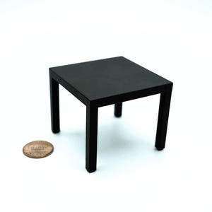 May include: A small, square black table with four legs. The table is made of a solid material and has a matte finish. A penny is next to the table for size comparison. The table is likely a miniature or dollhouse furniture.