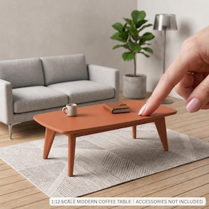 May include: A 1:12 scale modern coffee table in a matte terracotta finish with four tapered legs. The table surface holds a small beige ceramic mug and a brown rectangular notebook. Text on the image reads 1:12 SCALE MODERN COFFEE TABLE.