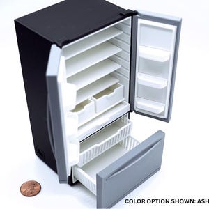 May include: A miniature black refrigerator with open doors revealing white interior shelves, drawers, and door bins. The bottom freezer drawer is pulled out, showing a gray front panel. A copper penny is placed next to the unit for scale. Text reads: COLOR OPTION SHOWN: ASH GRAY.