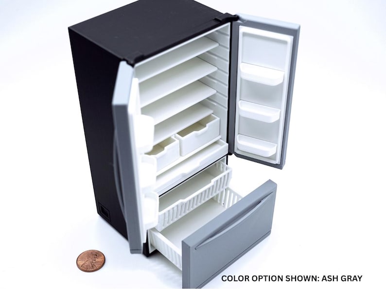 May include: A miniature black refrigerator with open doors revealing white interior shelves, drawers, and door bins. The bottom freezer drawer is pulled out, showing a gray front panel. A copper penny is placed next to the unit for scale. Text reads: COLOR OPTION SHOWN: ASH GRAY.