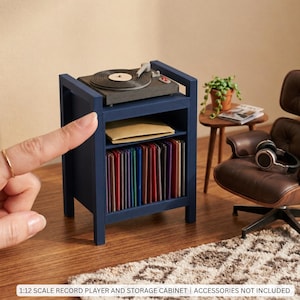 May include: A 1:12 scale miniature dark blue wooden record player storage cabinet featuring a top-mounted turntable, a middle shelf with a tan envelope, and a bottom shelf filled with colorful vinyl records. Text reads: 1:12 SCALE RECORD PLAYER AND STORAGE CABINET | ACCESSORIES NOT INCLUDED.