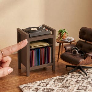 Pu&ograve; includere: Un supporto in miniatura in legno marrone per giradischi con un piatto sulla parte superiore, un ripiano centrale con una busta beige e un ripiano inferiore pieno di una collezione di dischi in vinile colorati, accanto a una poltrona marrone.