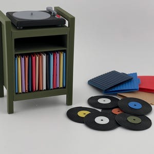 Pu&ograve; includere: Un supporto in miniatura per giradischi color verde oliva con un giradischi nero sopra e uno scaffale pieno di copertine colorate per dischi in vinile. Diversi dischi in vinile neri e altre copertine colorate sono posizionati accanto.