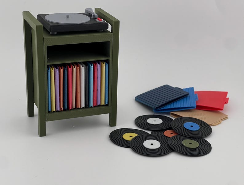 Pu&ograve; includere: Un supporto in miniatura per giradischi color verde oliva con un giradischi nero sopra e uno scaffale pieno di copertine colorate per dischi in vinile. Diversi dischi in vinile neri e altre copertine colorate sono posizionati accanto.