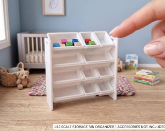 May include: A white 1:12 scale storage bin organiser featuring three horizontal rows of angled compartments. The unit is constructed with side panels and multiple removable white plastic bins. The text 1:12 SCALE STORAGE BIN ORGANISER | ACCESSORIES NOT INCLUDED is printed at the bottom.