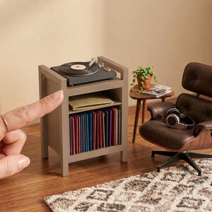 Pu&ograve; includere: Un supporto in miniatura grigio per giradischi con un piatto rotante sulla parte superiore, un ripiano centrale con una busta beige e un ripiano inferiore pieno di dischi in vinile multicolore, accanto a una poltrona marrone.