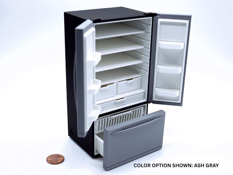 May include: A miniature black refrigerator with open doors revealing white interior shelves, drawers, and door bins. A gray freezer drawer is pulled open at the bottom. A copper penny is placed next to the unit for scale. Text reads: COLOR OPTION SHOWN: ASH GRAY.