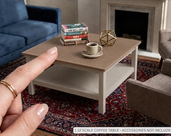 May include: A 1:12 scale square coffee table with a light brown textured top and a white frame. The table features a lower shelf and is displayed with miniature books, a small coffee cup on a saucer, and a gold geometric wire sculpture.