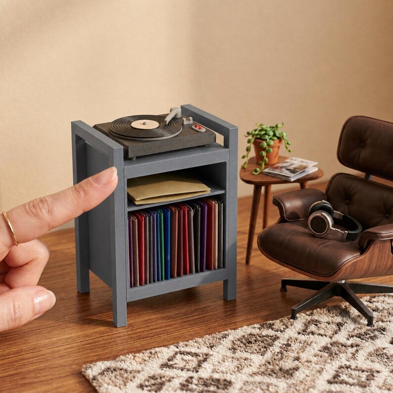 Pu&ograve; includere: Un mobiletto in miniatura in legno grigio con un giradischi sulla parte superiore, un ripiano centrale aperto con una busta beige e un ripiano inferiore pieno di una fila di dischi in vinile colorati, accanto a una poltrona marrone.