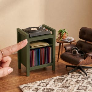Pu&ograve; includere: Un supporto in miniatura per giradischi in legno verde oliva con un piatto rotante sulla parte superiore, un ripiano centrale con una busta beige e un ripiano inferiore pieno di una collezione di dischi in vinile colorati, accanto a una poltrona marrone.