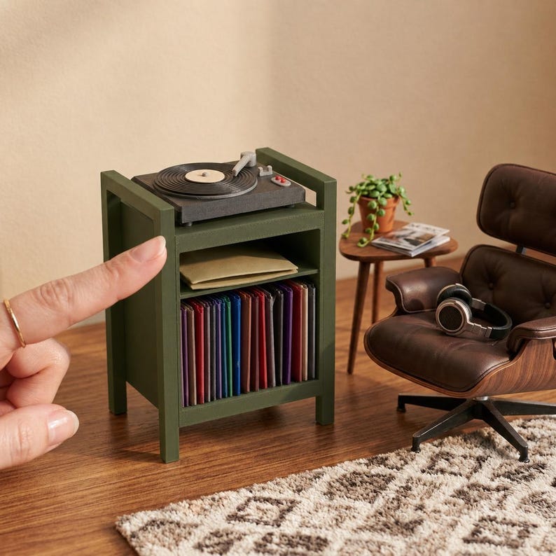 Pu&ograve; includere: Un supporto in miniatura per giradischi in legno verde oliva con un piatto rotante sulla parte superiore, un ripiano centrale con una busta beige e un ripiano inferiore pieno di una collezione di dischi in vinile colorati, accanto a una poltrona marrone.