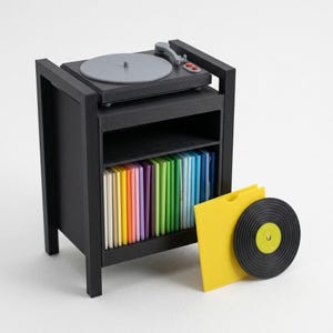 Pu&ograve; includere: Un supporto per giradischi in miniatura nero con un piatto grigio sulla parte superiore e uno scaffale pieno di una collezione di dischi in vinile color arcobaleno. Una custodia gialla e un disco in vinile nero sono posti davanti.