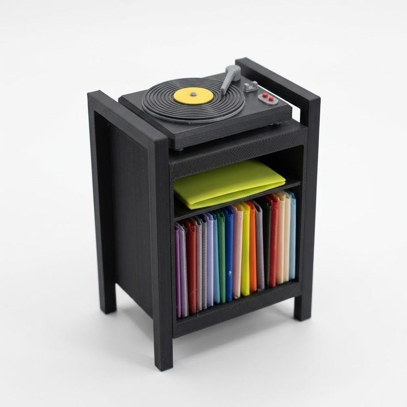 Pu&ograve; includere: Un supporto per giradischi in miniatura nero con un piatto montato superiormente con centro giallo, un ripiano centrale che sostiene un quadrato giallo brillante e un ripiano inferiore riempito con una fila di sottili custodie per dischi verticali multicolori.