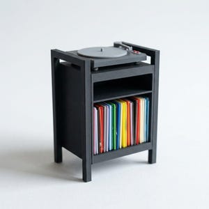 Pu&ograve; includere: Un supporto per giradischi in miniatura di colore nero con un piatto grigio sulla parte superiore e un ripiano aperto sottostante, riempito con una fila di sottili dischi in vinile disposti verticalmente in un arcobaleno di colori.