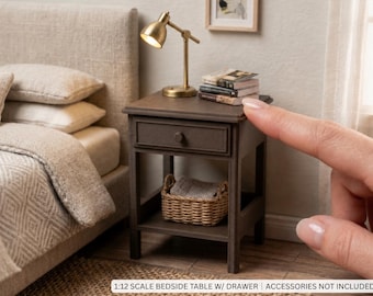 May include: A 1:12 scale brown wooden bedside table featuring a single drawer with a small round knob and an open lower shelf. The table surface holds a miniature brass-coloured lamp and a small stack of books, with a woven basket on the shelf.