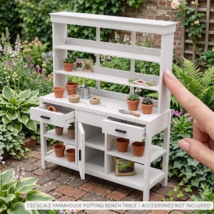 May include: A white 1:12 scale farmhouse potting bench table featuring a hutch with three shelves, two drawers with black handles, a cabinet door, and open lower shelving. The unit is displayed with miniature terracotta pots and garden tools.
