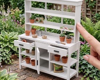 Può includere: Un tavolo da giardinaggio bianco in scala 1:12 con una credenza a tre ripiani, due cassetti con maniglie nere, un'anta e ripiani inferiori aperti, esposto con vasi in terracotta in miniatura e piccoli attrezzi da giardinaggio.