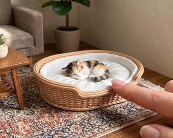 May include: A 1:12 scale wicker pet bed featuring a round, woven light brown frame and a soft, white tufted cushion. The bed contains a small, sleeping calico kitten figurine. Text on the image reads: 1:12 SCALE WICKER PET BED | CAT NOT INCLUDED.