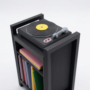 Pu&ograve; includere: Un giradischi in miniatura nero &egrave; posizionato sopra un mobiletto nero. Il piatto presenta un disco in vinile nero con un'etichetta centrale gialla, un braccio grigio e due pulsanti rossi. Sotto, lo scaffale contiene libri colorati.