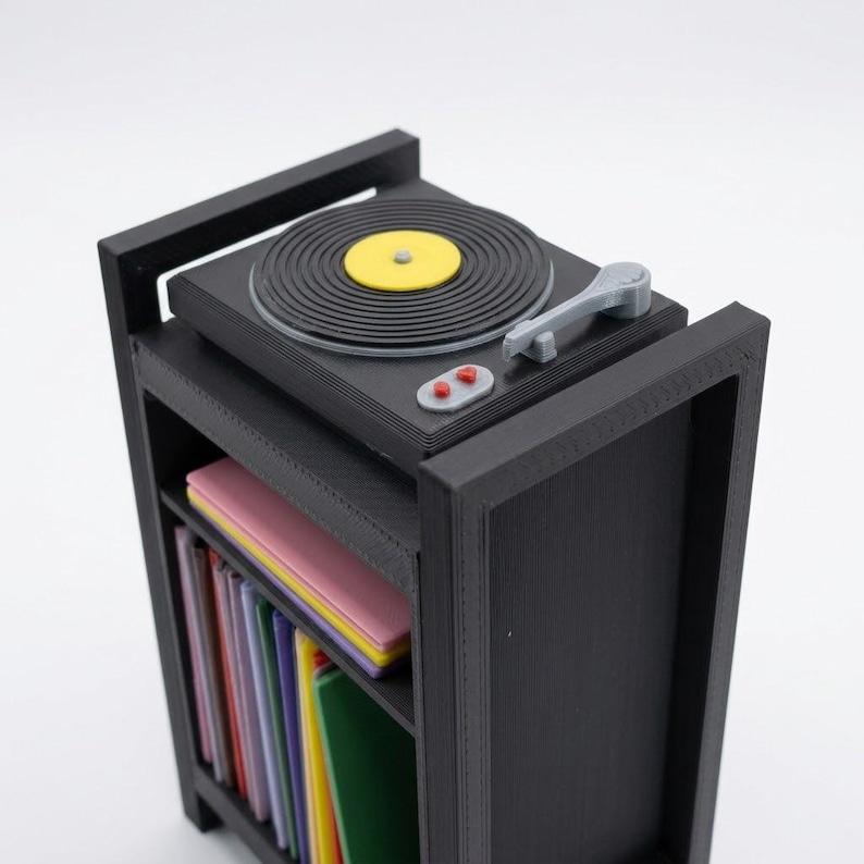 Pu&ograve; includere: Un giradischi in miniatura nero &egrave; posizionato sopra un mobiletto nero. Il piatto presenta un disco in vinile nero con un'etichetta centrale gialla, un braccio grigio e due pulsanti rossi. Sotto, lo scaffale contiene libri colorati.