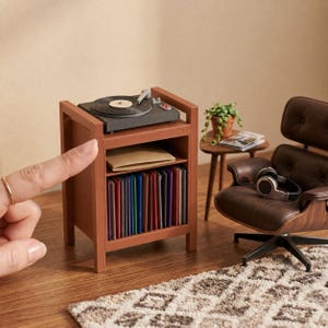 Pu&ograve; includere: Un supporto in miniatura in legno per giradischi con un giradischi nero sopra, un ripiano centrale con una busta beige e un ripiano inferiore pieno di dischi in vinile colorati, accanto a una poltrona marrone con cuffie.