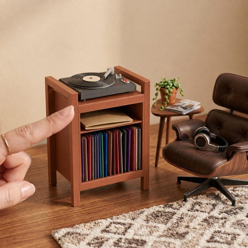 Pu&ograve; includere: Un supporto in miniatura in legno per giradischi con un giradischi nero sopra, un ripiano centrale con una busta beige e un ripiano inferiore pieno di dischi in vinile colorati, accanto a una poltrona marrone con cuffie.