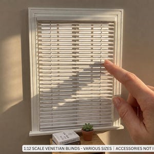 May include: A white miniature window frame with horizontal Venetian blinds. The frame features a classic molding design. Text at the bottom reads 1:12 SCALE VENETIAN BLINDS - VARIOUS SIZES | ACCESSORIES NOT INCLUDED. The blinds are closed.