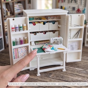 May include: A white miniature craft armoire featuring a fold-down desk, multiple storage shelves, and drawers. The unit contains small bottles of paint, spools of thread, a miniature sewing machine, fabric scraps, and tools. Text reads 1:12 SCALE CRAFT ARMOIRE W/ FOLD DOWN DESK | ACCESSORIES NOT INCLUDED.