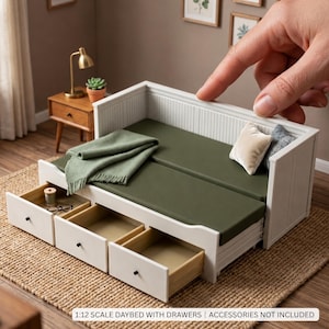 May include: A white miniature daybed with three pull-out drawers, featuring a dark olive green mattress, a folded green throw blanket, and two small decorative pillows. The frame has a vertical slat design. Text reads: 1:12 SCALE DAYBED WITH DRAWERS | ACCESSORIES NOT INCLUDED.