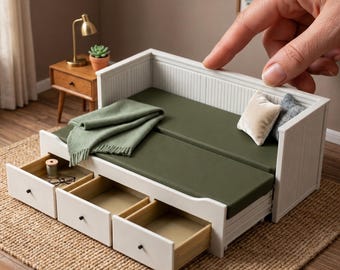 May include: A white miniature daybed with three pull-out drawers, featuring a dark olive green mattress, a folded green throw blanket, and two small decorative pillows. The frame has a vertical slat design. Text reads: 1:12 SCALE DAYBED WITH DRAWERS | ACCESSORIES NOT INCLUDED.